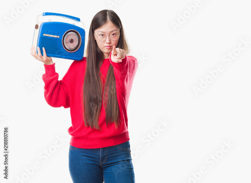 Young Chinese woman over isolated background holding vintage radio pointing with finger to the camera and to you, hand sign, positive and confident gesture from the front