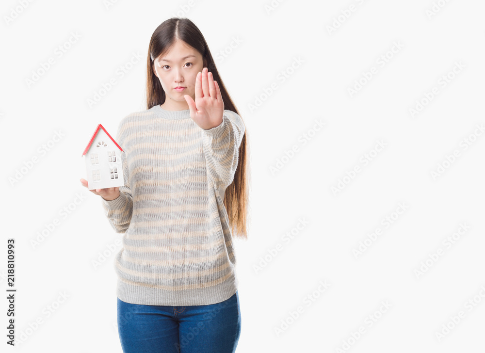 Young Chinese real state agent woman over isolated background holding house with open hand doing stop sign with serious and confident expression, defense gesture Young Chinese real state agent woman over isolated background holding house with open hand doing stop sign with serious and confident expression, defense gesture