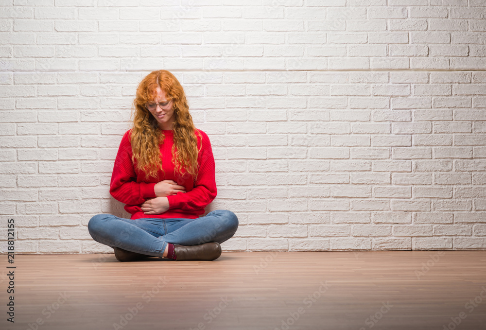 Young redhead woman sitting over brick wall with hand on stomach because indigestion, painful illness feeling unwell. Ache concept. Young redhead woman sitting over brick wall with hand on stomach because indigestion, painful illness feeling unwell. Ache concept.