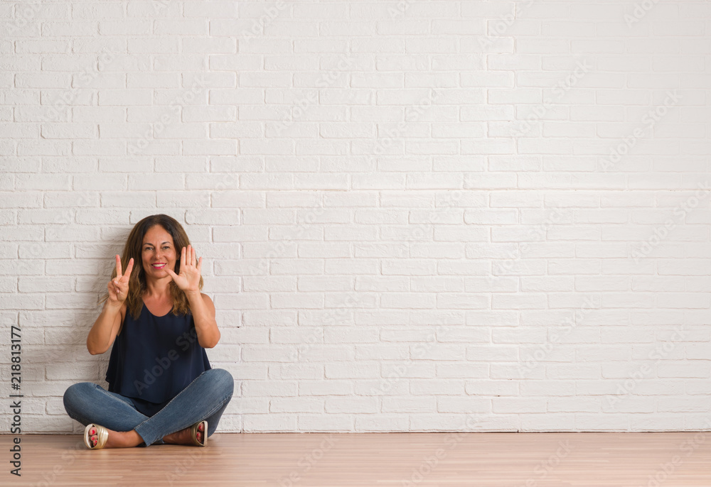 Middle age hispanic woman sitting on the floor over white brick wall showing and pointing up with fingers number seven while smiling confident and happy.