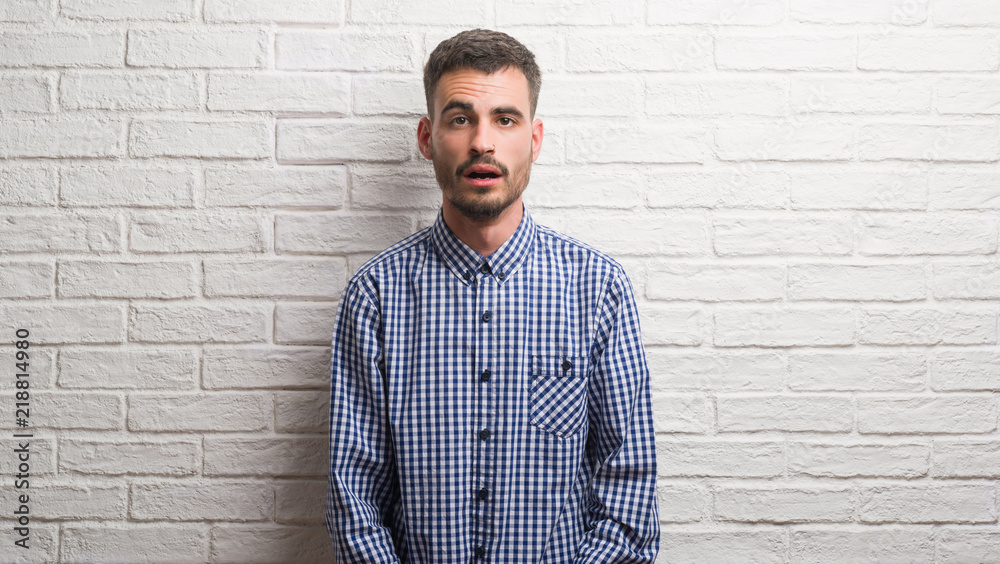 Young adult man standing over white brick wall scared in shock with a surprise face, afraid and excited with fear expression Young adult man standing over white brick wall scared in shock with a surprise face, afraid and excited with fear expression