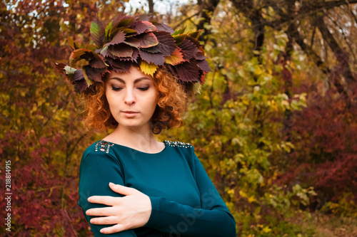 Redhead curly girl holding a wreath of grape leaves in a green dress. Young nymph in the wood. Copy space