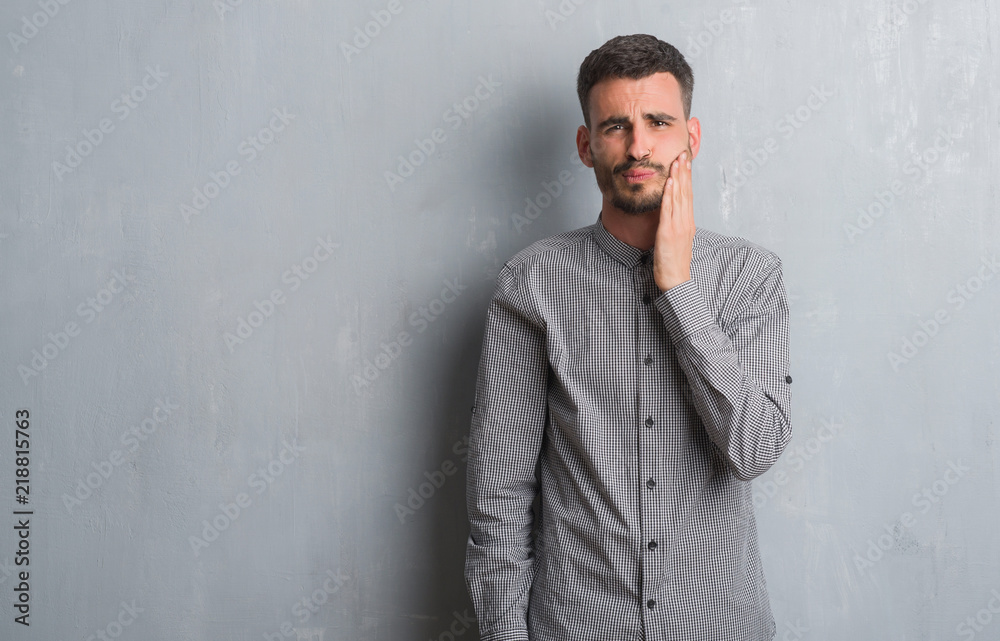 Young adult man standing over grey grunge wall touching mouth with hand with painful expression because of toothache or dental illness on teeth. Dentist concept.