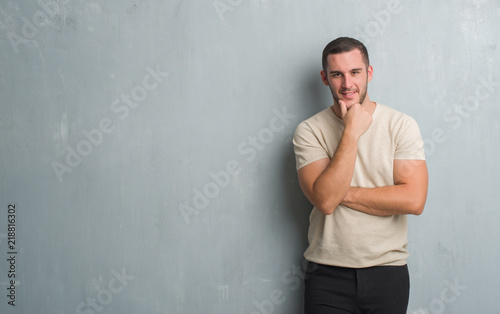 Young caucasian man over grey grunge wall looking confident at the camera with smile with crossed arms and hand raised on chin. Thinking positive.