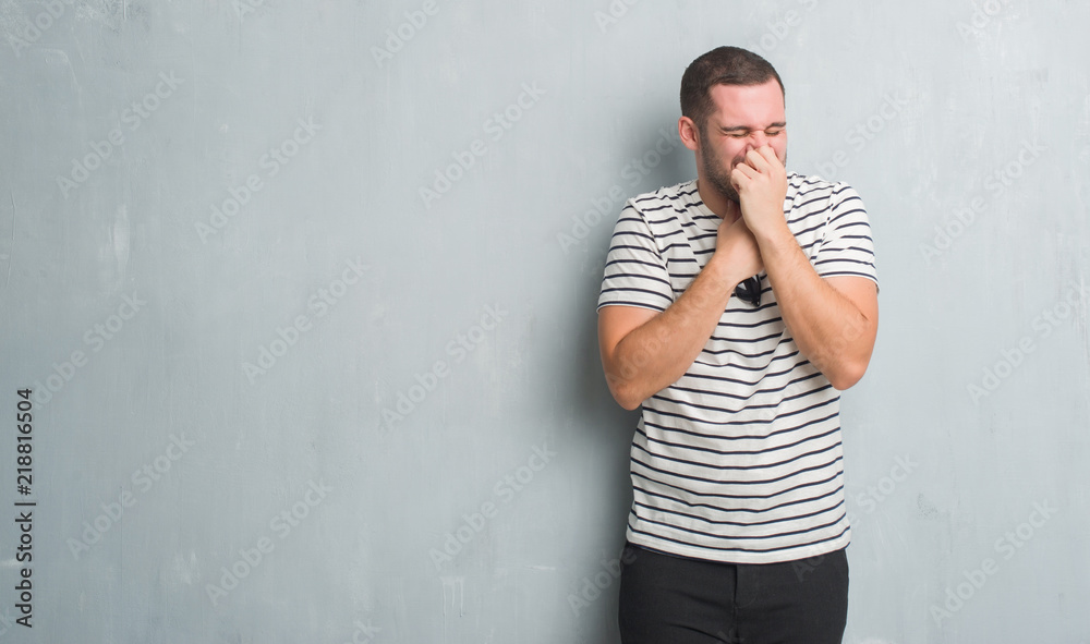 Young caucasian man over grey grunge wall smelling something stinky and disgusting, intolerable smell, holding breath with fingers on nose. Bad smells concept. Young caucasian man over grey grunge wall smelling something stinky and disgusting, intolerable smell, holding breath with fingers on nose. Bad smells concept.
