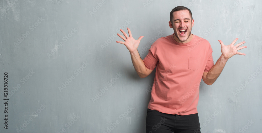 Young caucasian man over grey grunge wall celebrating mad and crazy for success with arms raised and closed eyes screaming excited. Winner concept Young caucasian man over grey grunge wall celebrating mad and crazy for success with arms raised and closed eyes screaming excited. Winner concept