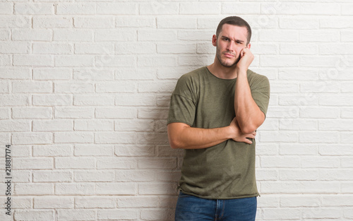 Young caucasian man standing over white brick wall thinking looking tired and bored with depression problems with crossed arms.