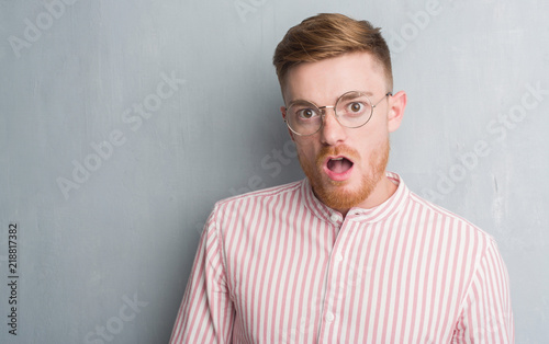 Young redhead man over grey grunge wall scared in shock with a surprise face, afraid and excited with fear expression