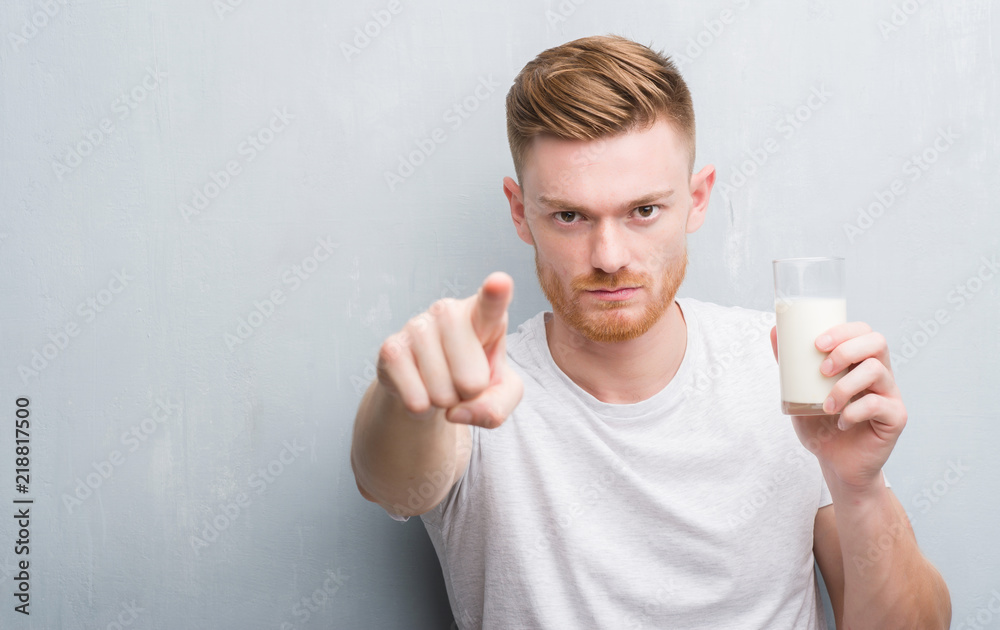 Young redhead man over grey grunge wall drinking a glass of milk pointing with finger to the camera and to you, hand sign, positive and confident gesture from the front Young redhead man over grey grunge wall drinking a glass of milk pointing with finger to the camera and to you, hand sign, positive and confident gesture from the front