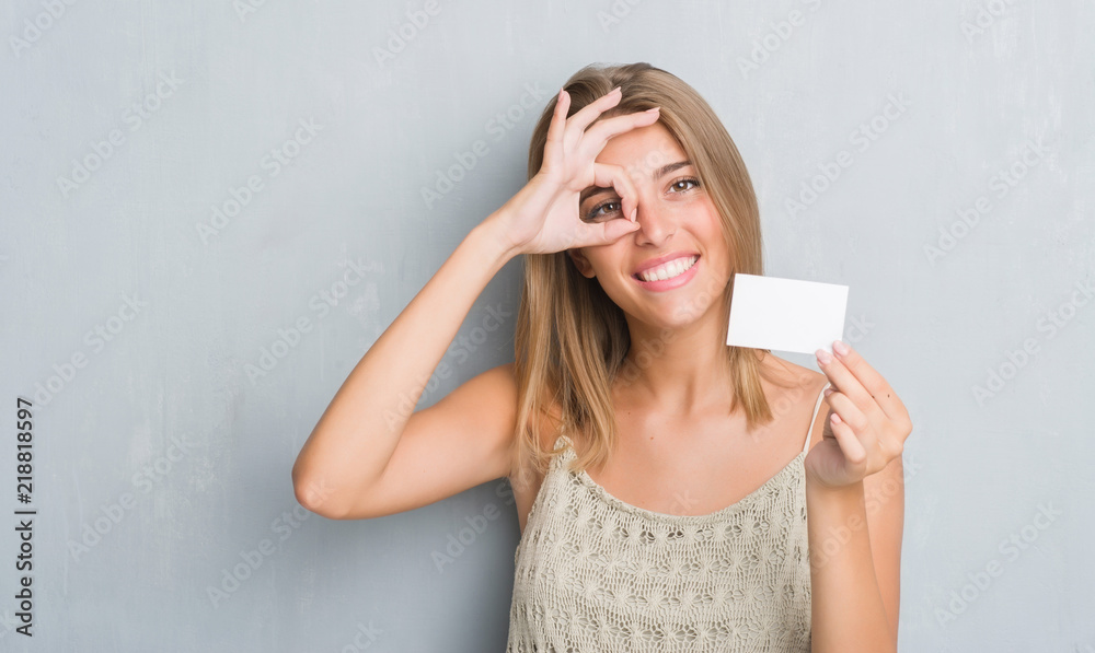 Beautiful young woman over grunge grey wall holding blank visit card with happy face smiling doing ok sign with hand on eye looking through fingers
