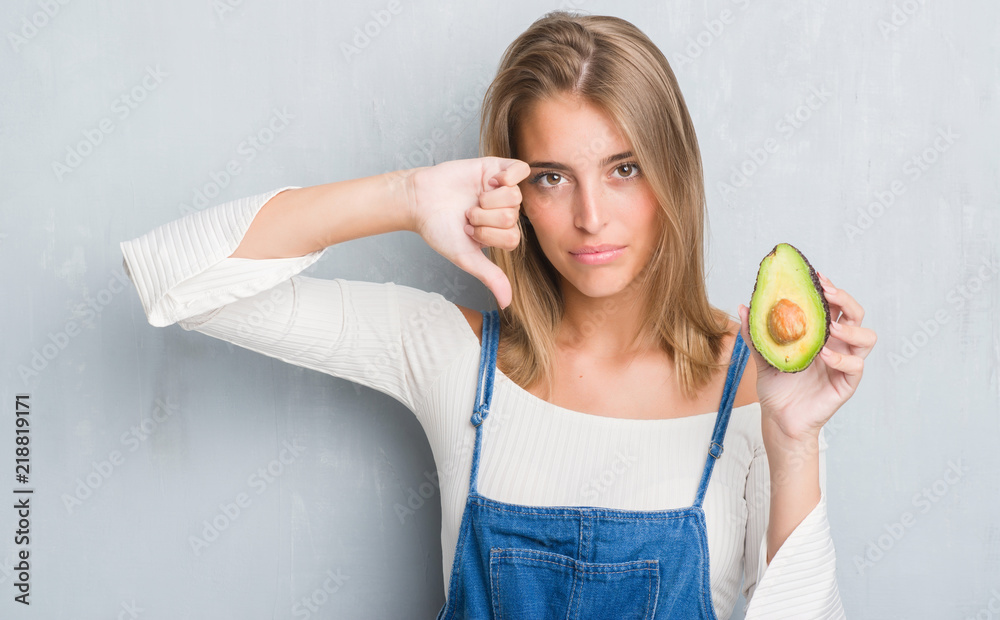 Beautiful young woman over grunge grey wall eating avocado with angry face, negative sign showing dislike with thumbs down, rejection concept Beautiful young woman over grunge grey wall eating avocado with angry face, negative sign showing dislike with thumbs down, rejection concept