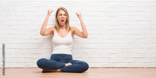 Beautiful young woman sitting on the floor at home angry and mad raising fist frustrated and furious while shouting with anger. Rage and aggressive concept.