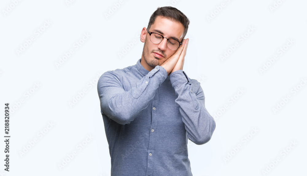 Handsome young elegant man wearing glasses sleeping tired dreaming and posing with hands together while smiling with closed eyes. Handsome young elegant man wearing glasses sleeping tired dreaming and posing with hands together while smiling with closed eyes.