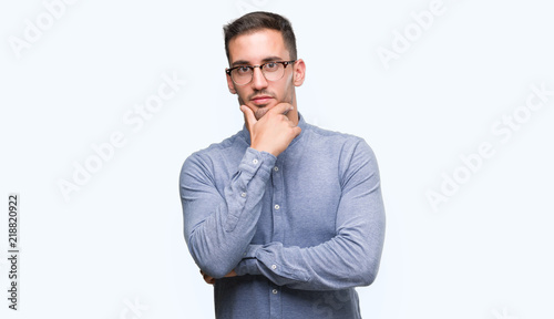 Handsome young elegant man wearing glasses looking confident at the camera with smile with crossed arms and hand raised on chin. Thinking positive.