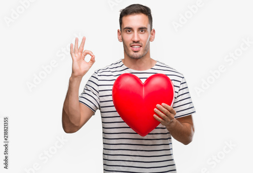 Handsome young man in love holding red heart doing ok sign with fingers, excellent symbol © Krakenimages.com