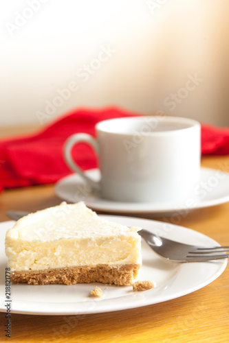 Homemade vanilla cheesecake on a white plate with a fork and a white cup and red cloth in the background
