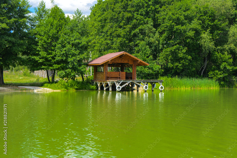 Obraz premium A beautiful image of a landscape from center of a river surrounded by trees and reeds on the shore against a blue sky in the clouds. Wooden gazebo on the beach. Reflection, water, tourist destination