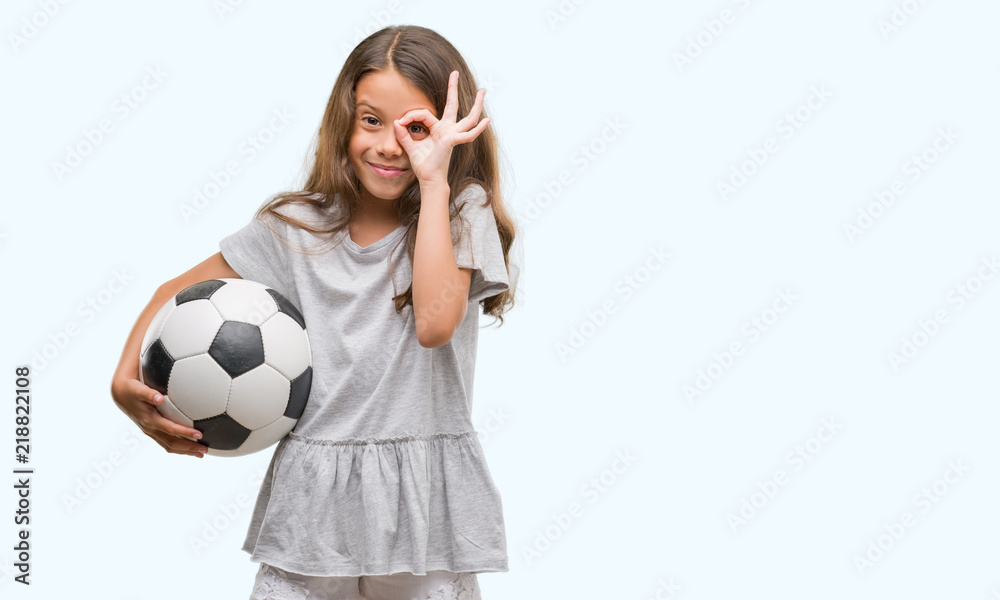 Brunette hispanic girl holding soccer football ball with happy face smiling doing ok sign with hand on eye looking through fingers Brunette hispanic girl holding soccer football ball with happy face smiling doing ok sign with hand on eye looking through fingers