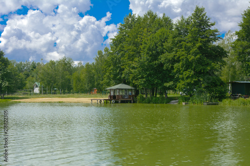 A beautiful image of a landscape from center of a river surrounded by trees and reeds on the shore against a blue sky in the clouds. Wooden gazebo on the beach. Reflection, water, tourist destination