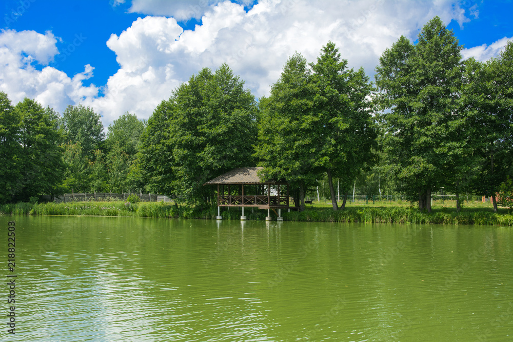 A beautiful image of a landscape from center of a river surrounded by trees and reeds on the shore against a blue sky in the clouds. Wooden gazebo on the beach. Reflection, water, tourist destination