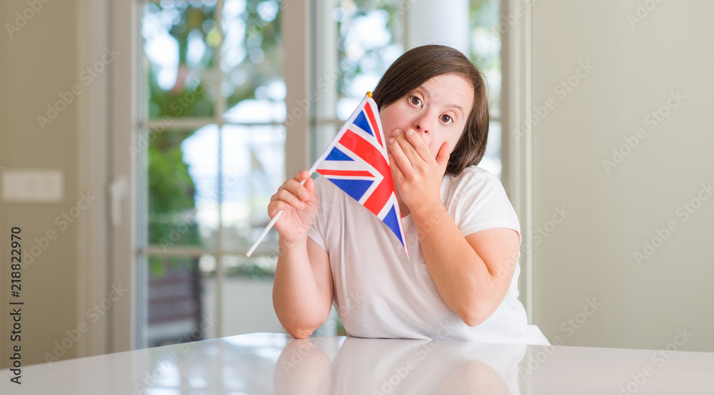 Down syndrome woman at home holding flag of uk cover mouth with hand shocked with shame for mistake, expression of fear, scared in silence, secret concept