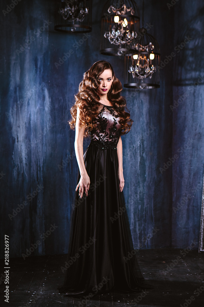 Young woman with long curly hair and makeup in evening long luxury dress, posing in a dark interior room. fashion beauty portrait