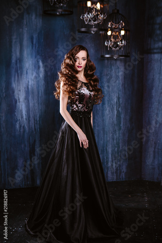 Young woman with long curly hair and makeup in evening long luxury dress  posing in a dark interior room. fashion beauty portrait