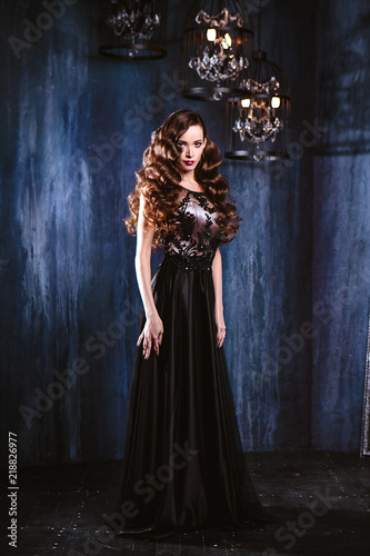 Young woman with long curly hair and makeup in evening long luxury dress, posing in a dark interior room. fashion beauty portrait © cherry_d