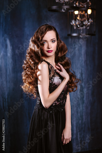 Young woman with long curly hair and makeup in evening long luxury dress, posing in a dark interior room. fashion beauty portrait