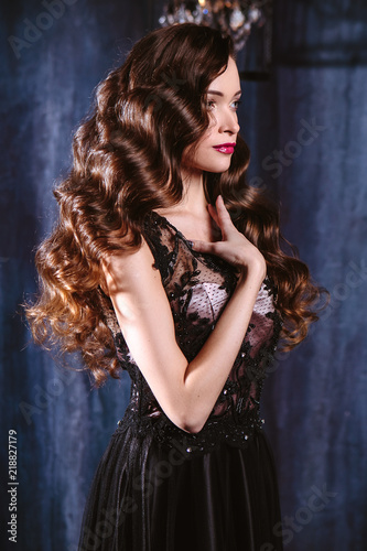 Young woman with long curly hair and makeup in evening long luxury dress, posing in a dark interior room. fashion beauty portrait