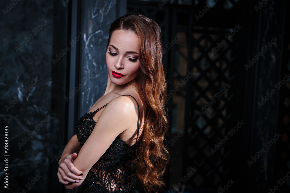 Young woman with long curly hair and makeup in evening long luxury dress, posing in a dark interior room. fashion beauty portrait