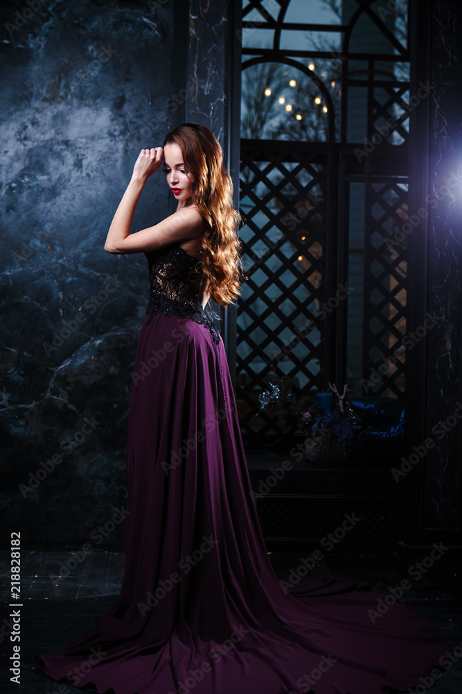 Young woman with long curly hair and makeup in evening long luxury dress, posing in a dark interior room. fashion beauty portrait
