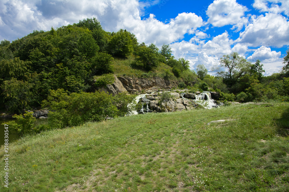 The waterfall on the river flows through and over the rocks covered with lichen and moss against a background of green vegetation and a blue sky.