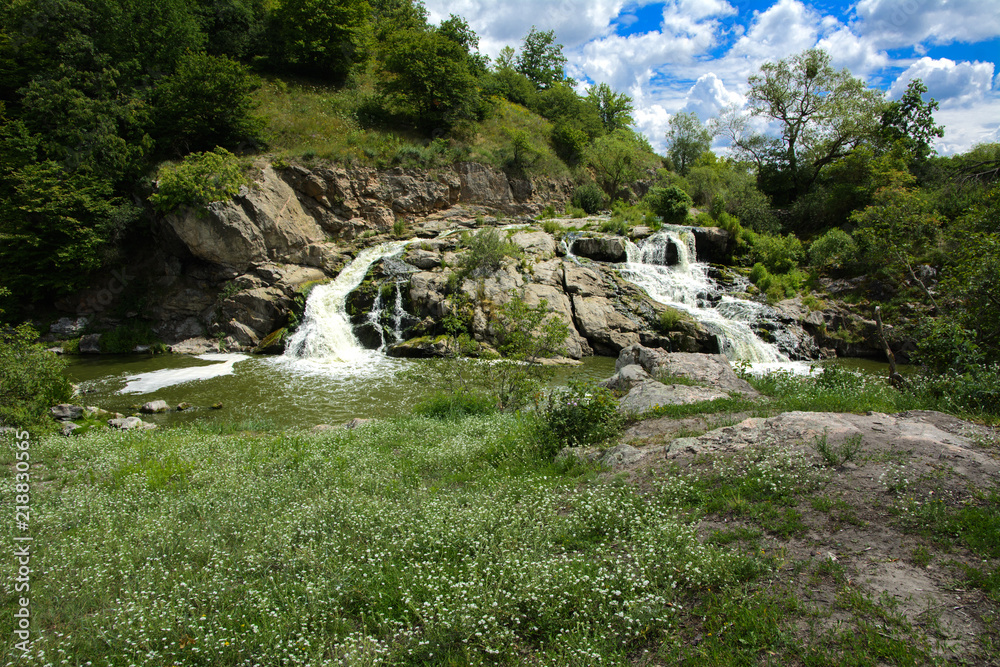 The waterfall on the river flows through and over the rocks covered with lichen and moss against a background of green vegetation and a blue sky.