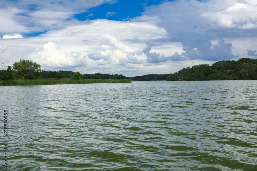 A beautiful image of landscape from the center of the river, surrounded by trees and reeds on the shore and distant horizon against the blue sky in clouds. Reflection, water, tourist destination A beautiful image of landscape from the center of the river, surrounded by trees and reeds on the shore and distant horizon against the blue sky in clouds. Reflection, water, tourist destination