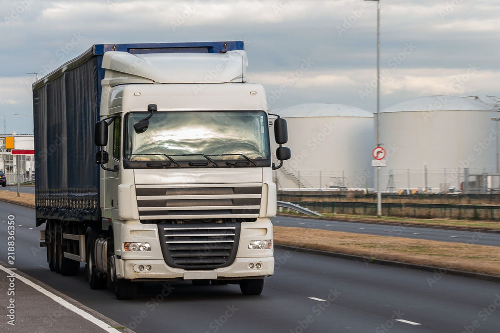 Naklejka premium Road transport. Large blue lorry in motion on the road with oil depot in the background