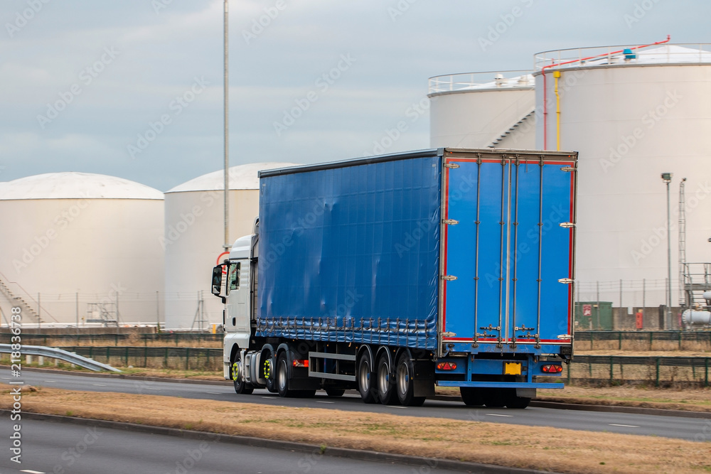 Road transport. Large blue lorry in motion on the road with oil depot in the background Road transport. Large blue lorry in motion on the road with oil depot in the background