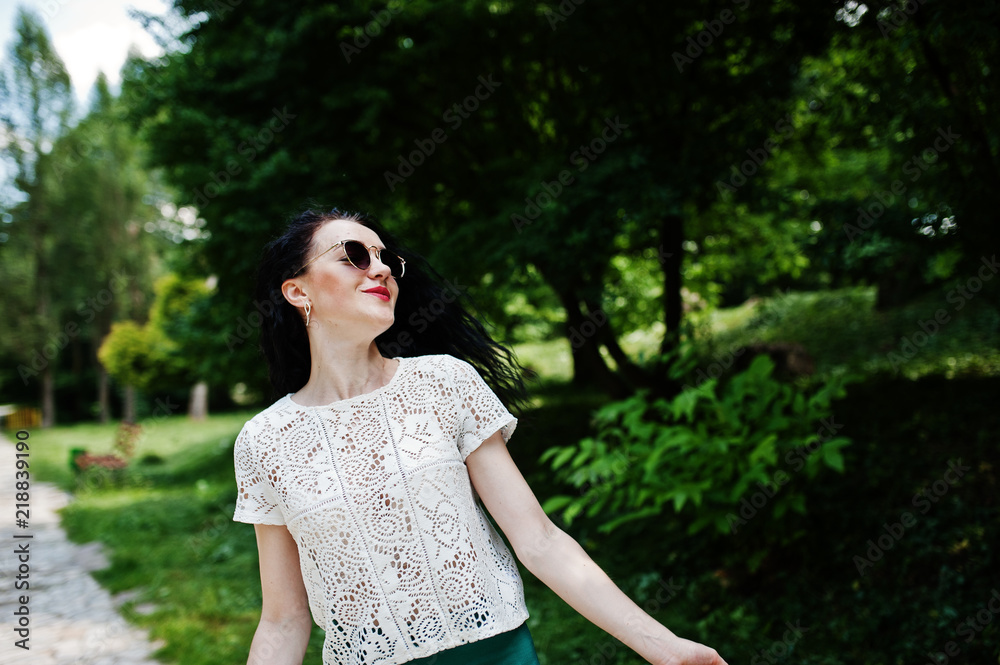 Brunette girl in green skirt and white blouse with sunglasses posed at park.