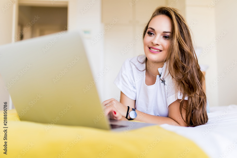 A smiling woman lying down the bed in front of her laptop with her legs raised slightly at home