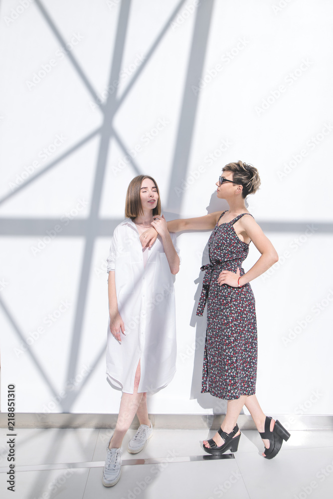 Fototapeta premium Two fashionable girlfriends stand in full swing at the background of a white wall with shadows. Portrait of two stylish attractive girls on the background of a white wall.