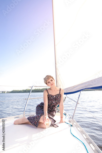 Attractive woman with short hair and a dress is seated on a sailboat with a glass of wine and resting. Beautiful woman floating on a yacht by sea on a summer sunny day. Rest on a yacht