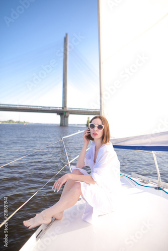 Attractive woman wearing glasses and white clothes poses sitting on a yacht on the background of a river and a bridge. Stylish young woman is having a rest, sailing on a river by boat.