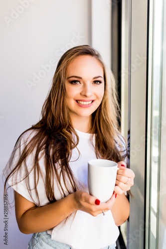Morning coffee is my daily routine. Young woman drink cup of morning coffee near windows at home looking at street. Morning mood.