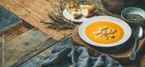 Fall warming pumpkin cream soup with croutons and seeds on board over rustic wooden background, copy space, wide composition. Autumn vegetarian, vegan, healthy comfort food concept