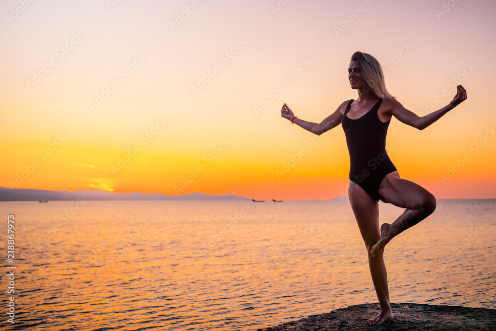Beautiful girl in black doing yoga on the pier by the sea, standing asana Vrikshasana at sunrise. Healthy lifestyle.