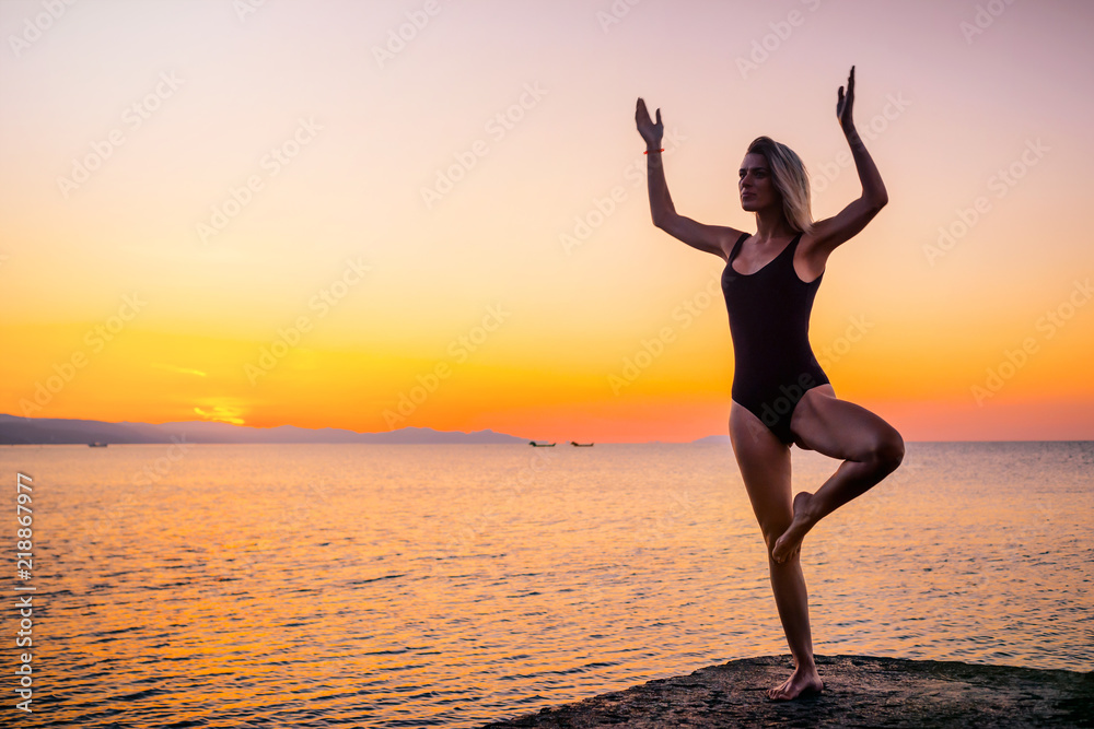Beautiful girl in black doing yoga on the pier by the sea, standing asana Vrikshasana at sunrise. Healthy lifestyle.