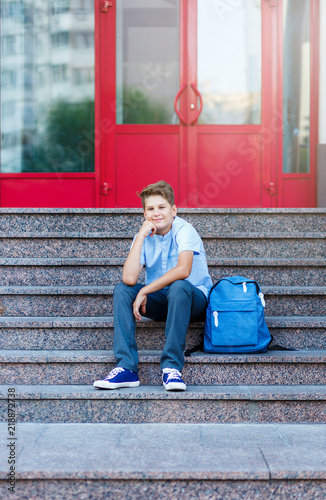 cute, young boy in blue shirt sits on the steps wit backpack in front of his schhool. Education, back to school concept photo