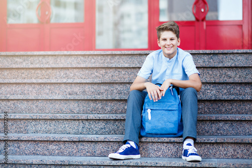 cute, young boy in blue shirt sits on the steps wit backpack in front of his schhool. Education, back to school concept photo