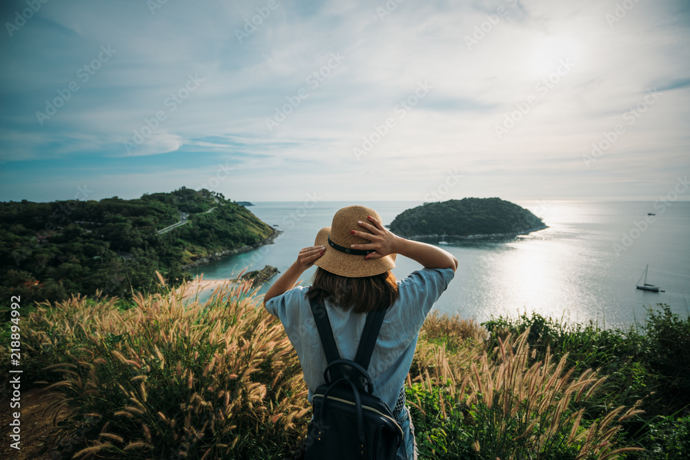 Fototapeta premium Young asian woman traveler with backpack and hat looking view at view point look sunset at Promthep Cape Phuket, Thailand.