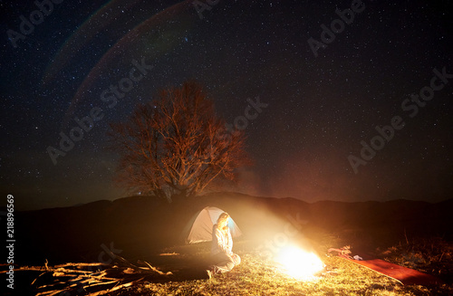 Camping night in mountains. Female tourist having a rest near burning bonfire under deep dark starry sky with brightly lit tent and silhouette of big tree in background. Tourism and travel concept.